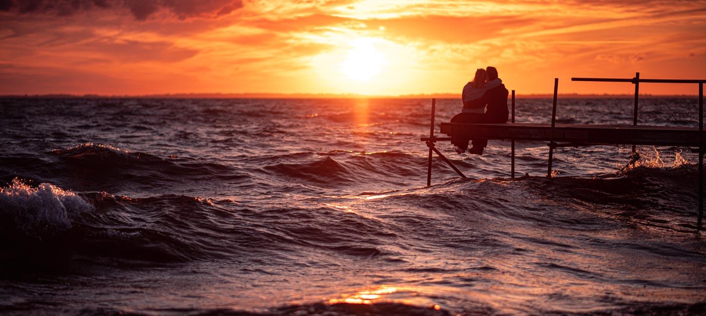 Et par sidder på en træbro over havet og iagttager en intens rød solnedgang.