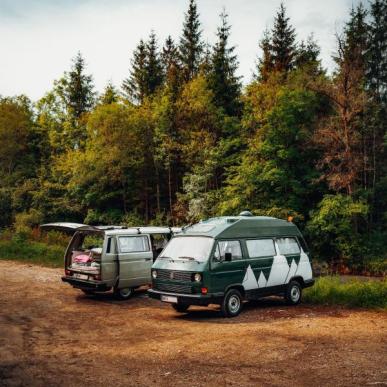 To autocamper vans holder parkeret i skovkanten, den ene med bagagesmækken og sidedøren åben. Høje nåletræer i baggrunden.