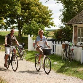 Et par cykler igennem idyllisk landskab med stråtagshus, hvidt stakit og grønne træer i baggrunden.