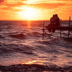 Et par sidder på en træbro over havet og iagttager en intens rød solnedgang.