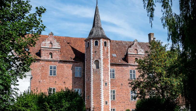 Herregården Harridslevgaard ses gennem flere træer. Gaarden er rød og har i midten et tårn med et helt spidst tag.