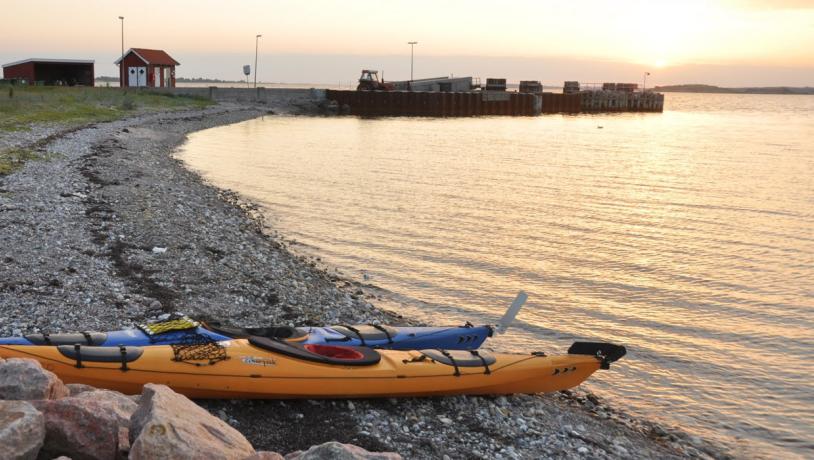 På billedet ses en strandbred. Den fylder hele venstre side af billedet og ender i baggrunden af billedet. Her står to små røde bygninger. Henover strandbredden er solen på vej ned. I højre side er havet. i forgrunden ligger en gul og en blå kajak på standen.