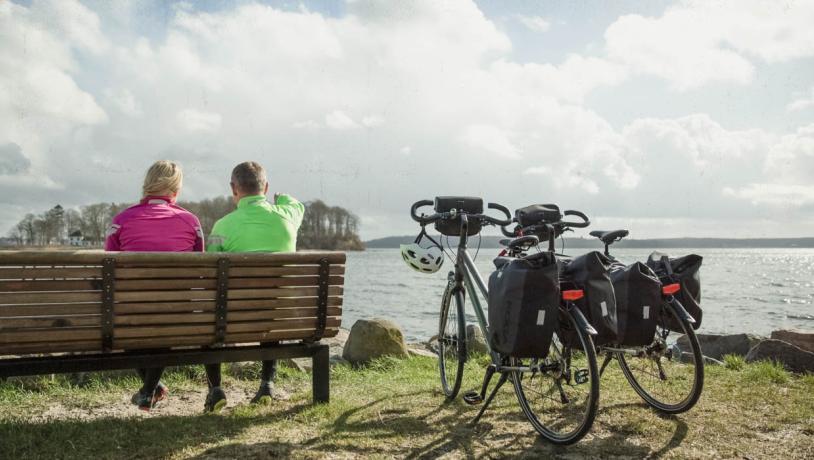 To cyklister i neon-arvede sportsjakker sidder med ryggen til på en bænk og ser ud over sundet. To cykler med cykeltasker på står parkeret ved siden af.