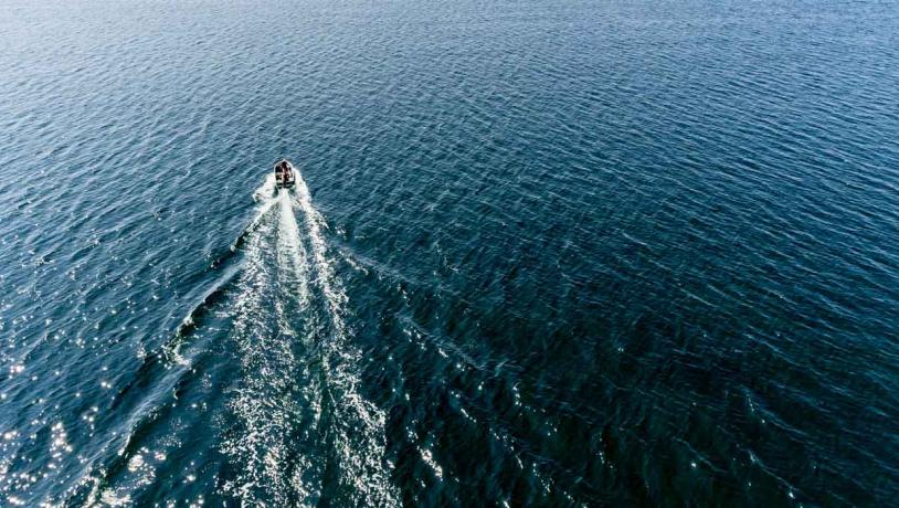 Dronebillede af en jolle motor båd sejler midt i det åbner hav. Bag den ses et sejlspor af hvidt havskum.
