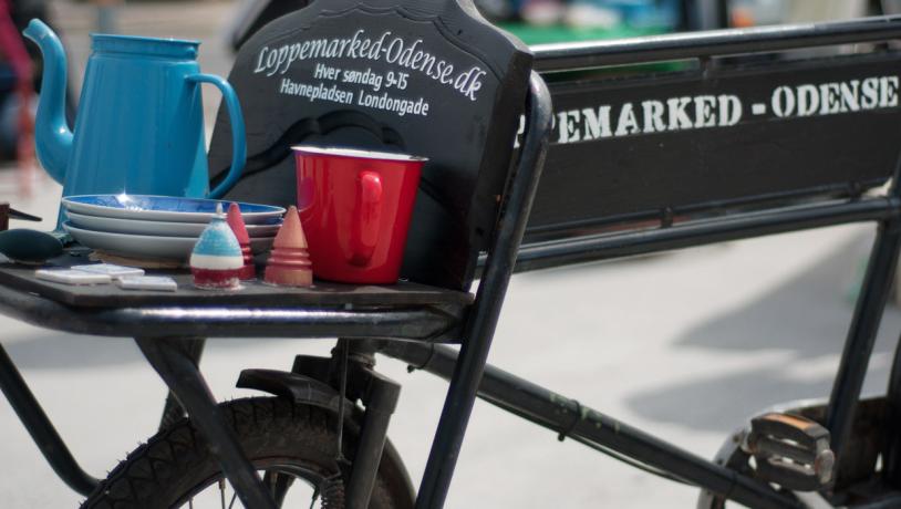 En nært billede af en sort gammel cykel. Cyklen har et lille lad foran med teksten "Loppemarked-Odense.dk" skrevet på. På ladet står en blå kande, par blå tallerkner og en rød kop.