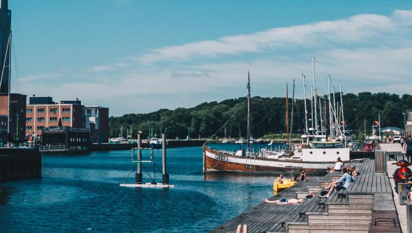 Mennesker soler sig på træplatformen ud til vandet ved Odense havn. Der er blå himmel og lidt skyer. 