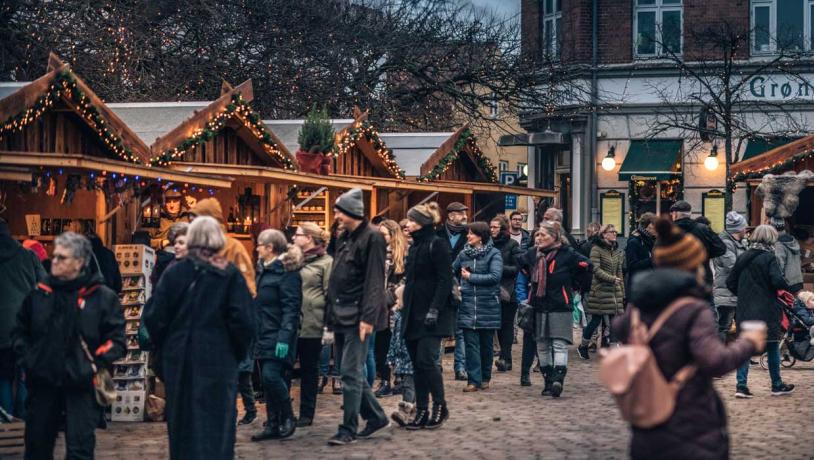 En brostensplads er fyldt med mennesker pakket ind i vintertøj. Rundt om pladsen står små træhytter pyntet med gran og julelys. Træhytterne har en åben front som giver adgang til de juleting de sælger.