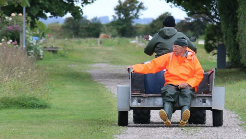 To personer på en havetraktor med trailer-lad. Manden, der kører traktoren, har ryggen til og er iklædt en grøn jakke. Den anden mand sidder bagpå på ladet med benene hængende ud over kanten i en orange jakke. De kører afsted på en grussti i grønne omgivelser.