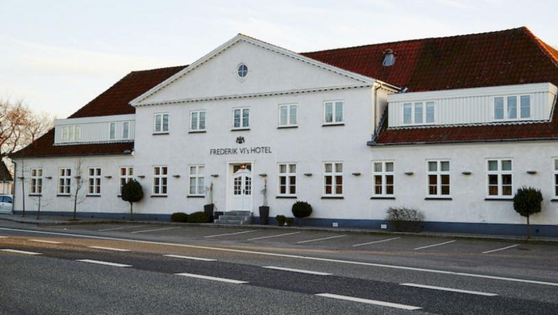 Frederik VI's hotel set ude fra vejen. Bred, hvid bygning med rødt tag ud mod asfaltvej med hvide striber.