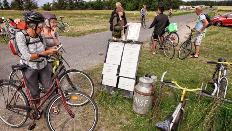 En flok mennesker er samlet omkring et cykeludlejningssted. To ældre kvinder står i forgrunden med cykler og cykelhjelm og læser et skilt.