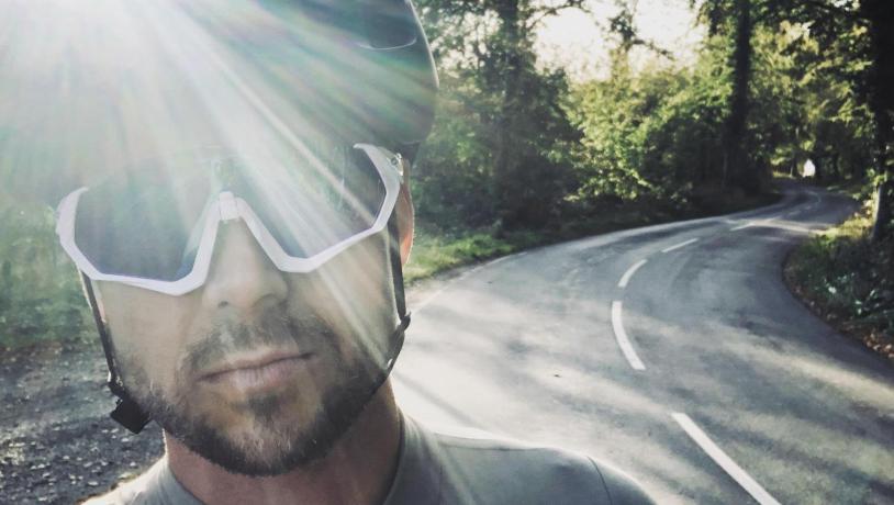 Portrætfoto af Bo Jessen med cykelhjelm og -briller oplyst af sollys og med fynsk landevej i baggrunden.