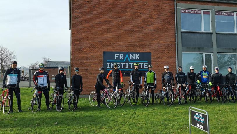 En flok cyklister står i fuldt cykel-outfit og med deres cykler i hånden på én lang række foran en rød bygning med et skilt med teksten "Frank Institute of Sports"