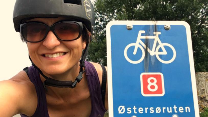 En kvinde i lilla tanktop, sorte solbriller og en sort cykelhjelm tager et selfie med et blå skilt. På skiltet er en hvid cykel og teksten "Østersøruten". i Bagrunden er en stort grantræ.