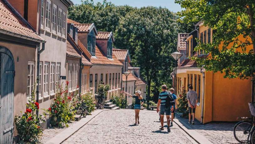 Gammel stræde med brosten og bindingsværkshuse i orange, røde og beige farver. En flok mennesker går tur i det gode vejr og kigger på husene.