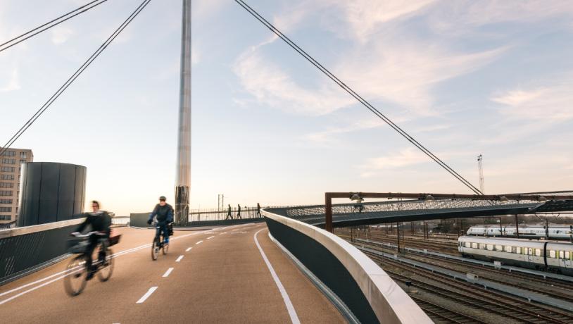 Byens Bro i Odense med to cyklister i fart på vej henover broen. Etagebygning i baggrunden, og togene nede på skinnerne til højre i billedet.