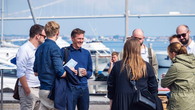 I forgrunden står otte mennesker i en cirkel og snakker sammen. I baggrunden ses en masse skibe der ligger i havn. I horisonten kan man se Lillebælt broen.