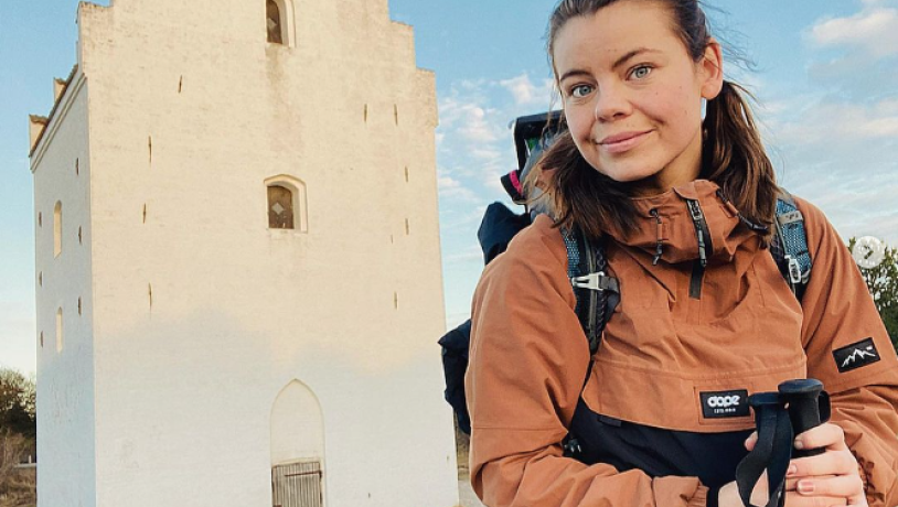 Influenseren Astrid Vendelbo står foran den tilsandede kirke. Hun har vandretøj og rygsæk på og stave i hænderne. 