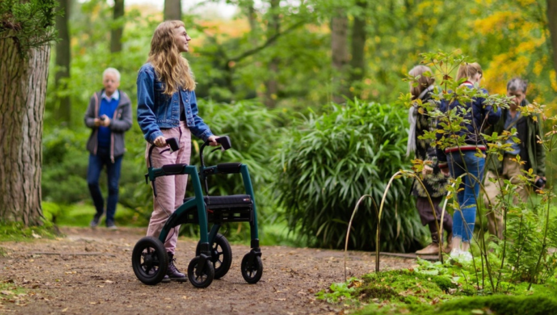 Ung pige med rollator står i grønne omgivelser med andre mennesker i baggrunden