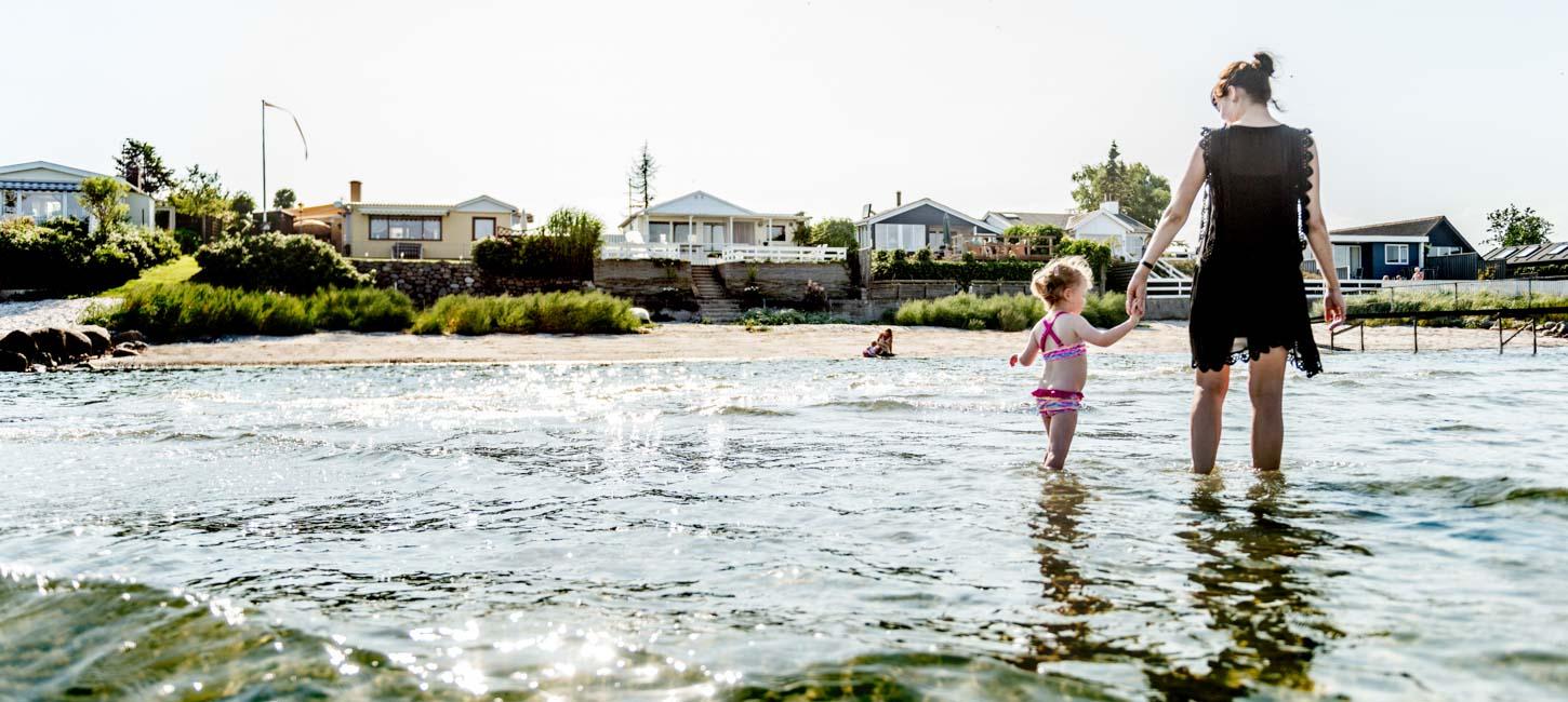 En mor går med en lille pige i hånden og sopper i vandet langs strandkanten.