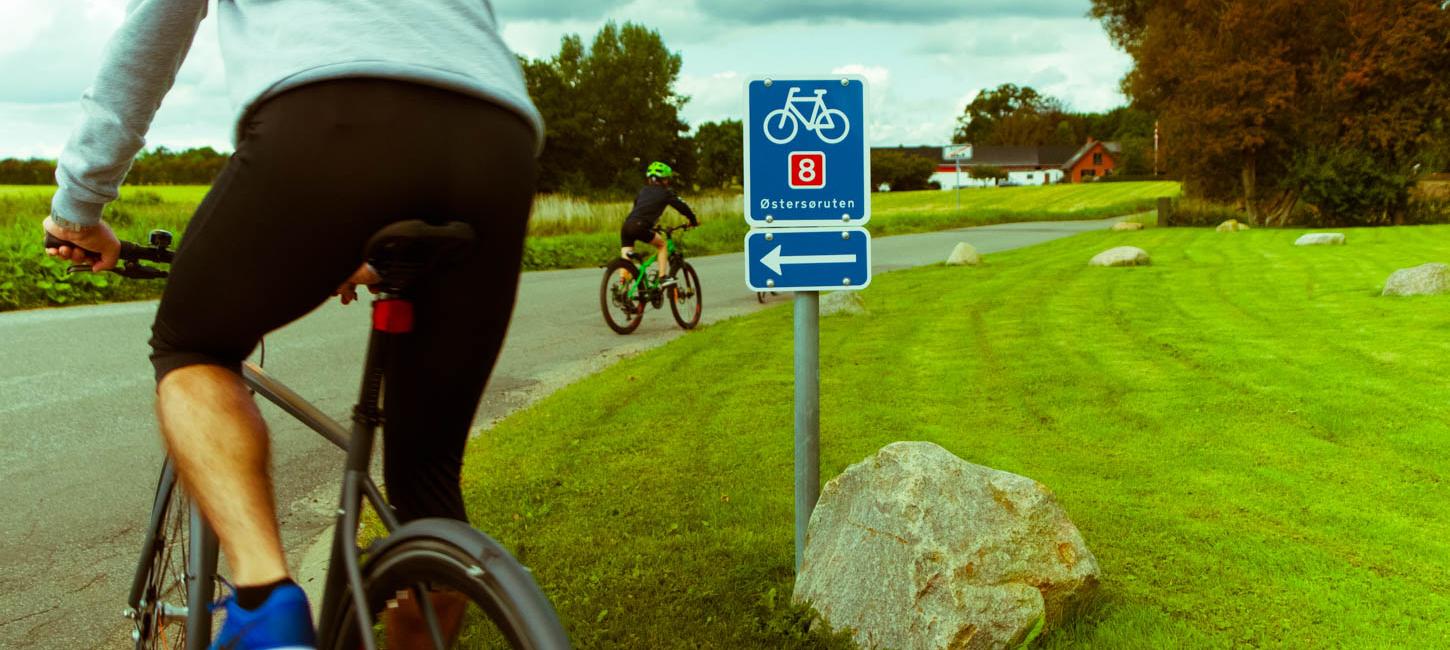 To cyklister kører afsted forbi blåt skilt med hvidt cykelikon, et rødt 8-tal og teksten Østersøruten.