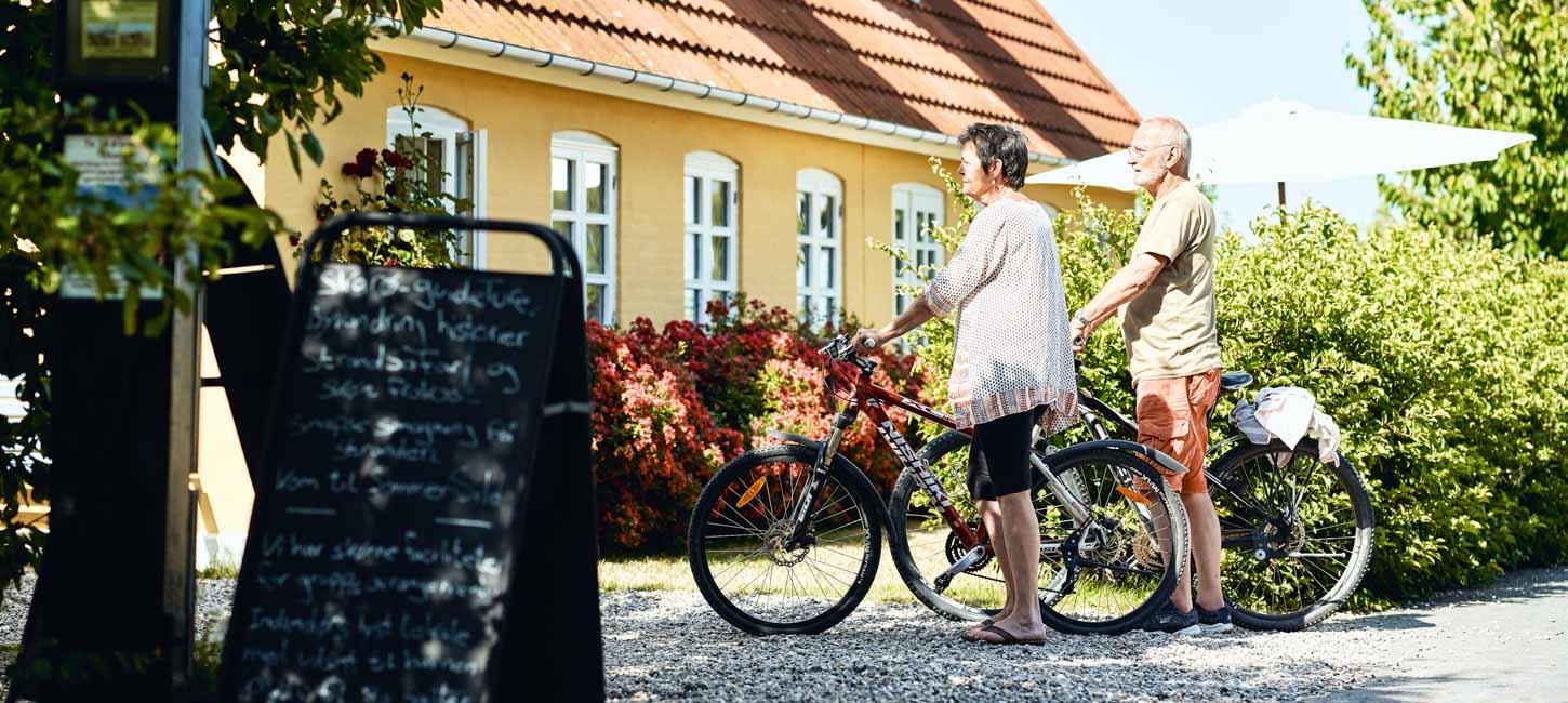 Mand og kvinde står afventende med deres cykler ved gult hus. 