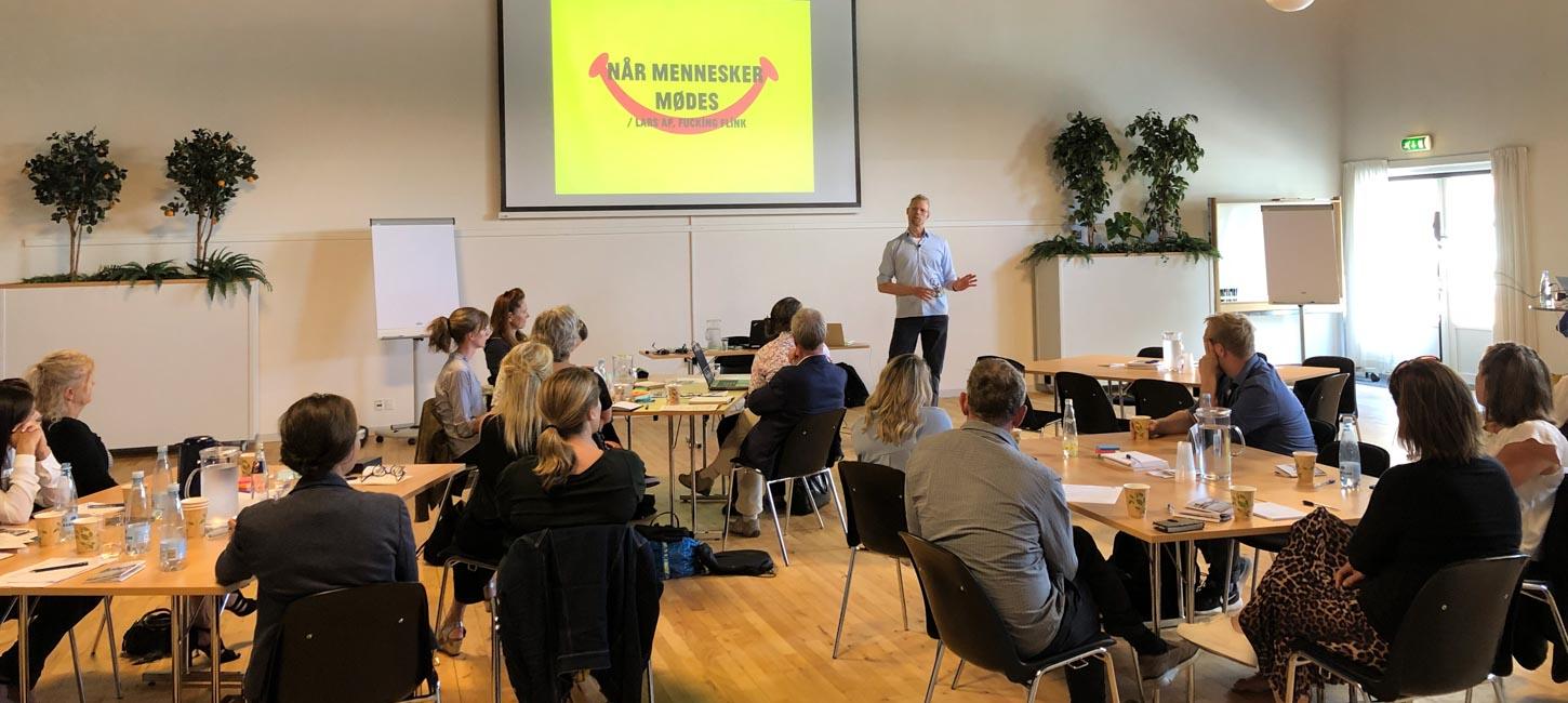 En del mennesker, siddende omkring borde med ryggen til, kigger på på oplægsholder, der er i gang med en PowerPoint-præsentation. 