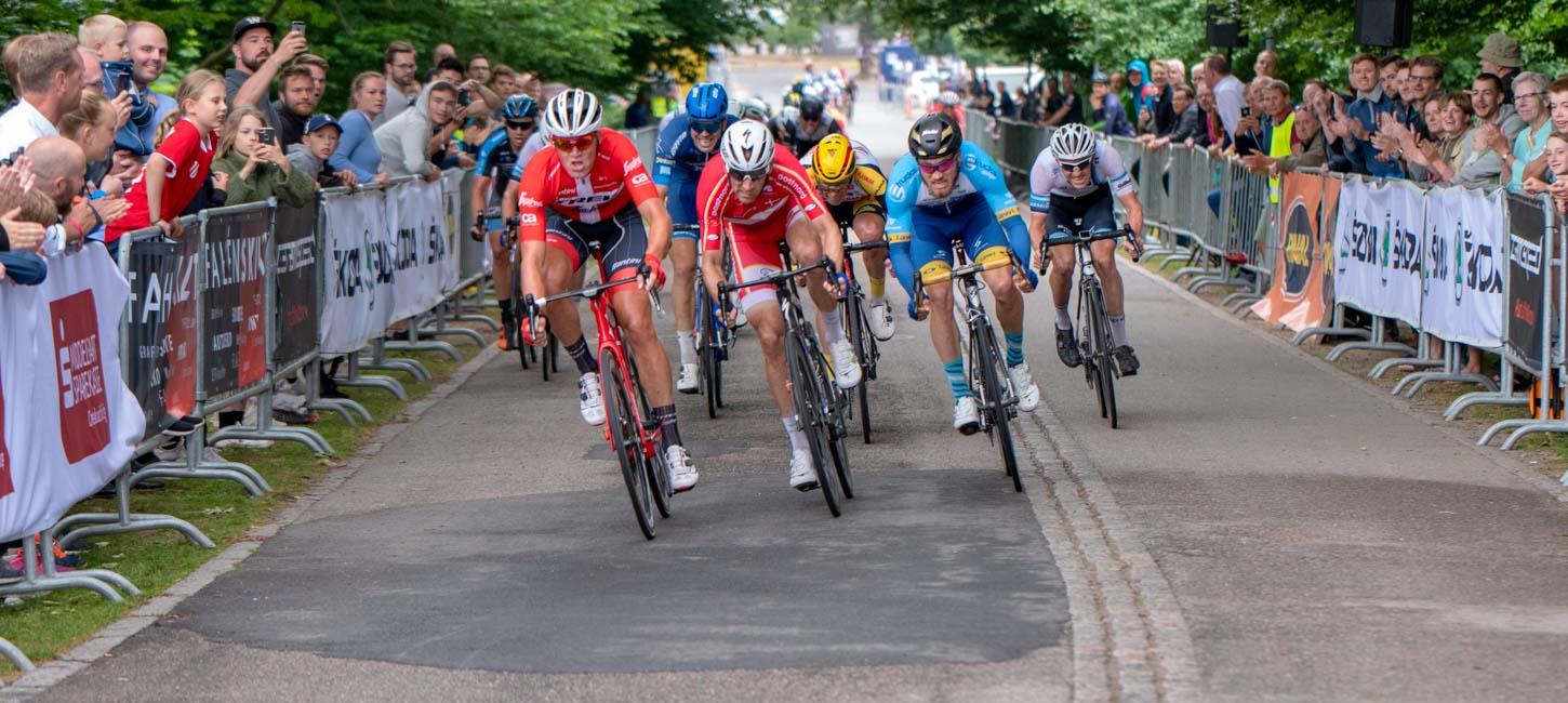 Cykelryttere på vej ind ad opløbsstrækningen i tæt løb. Tilskuere står bag hegn og hepper til begge sider.