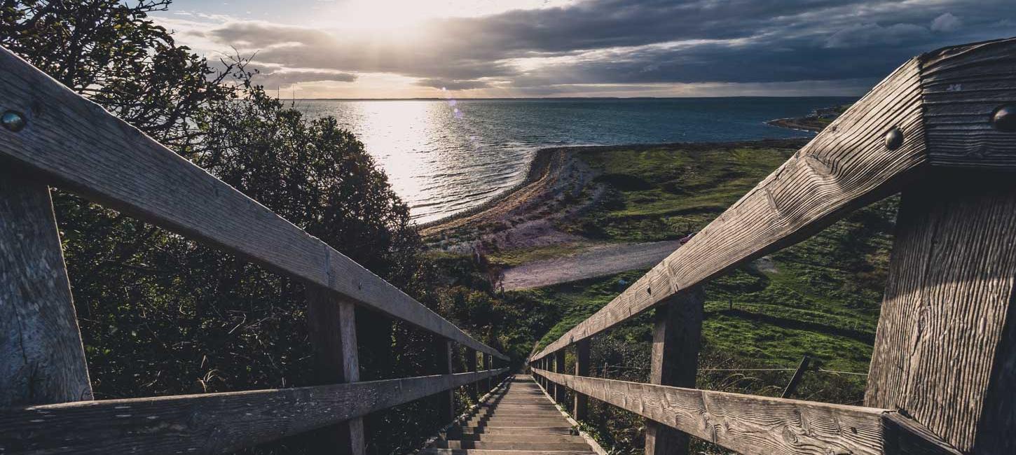 Udsigt fra toppen af lang trætrappe udover havet, som solen oplyser på dramatisk vis.