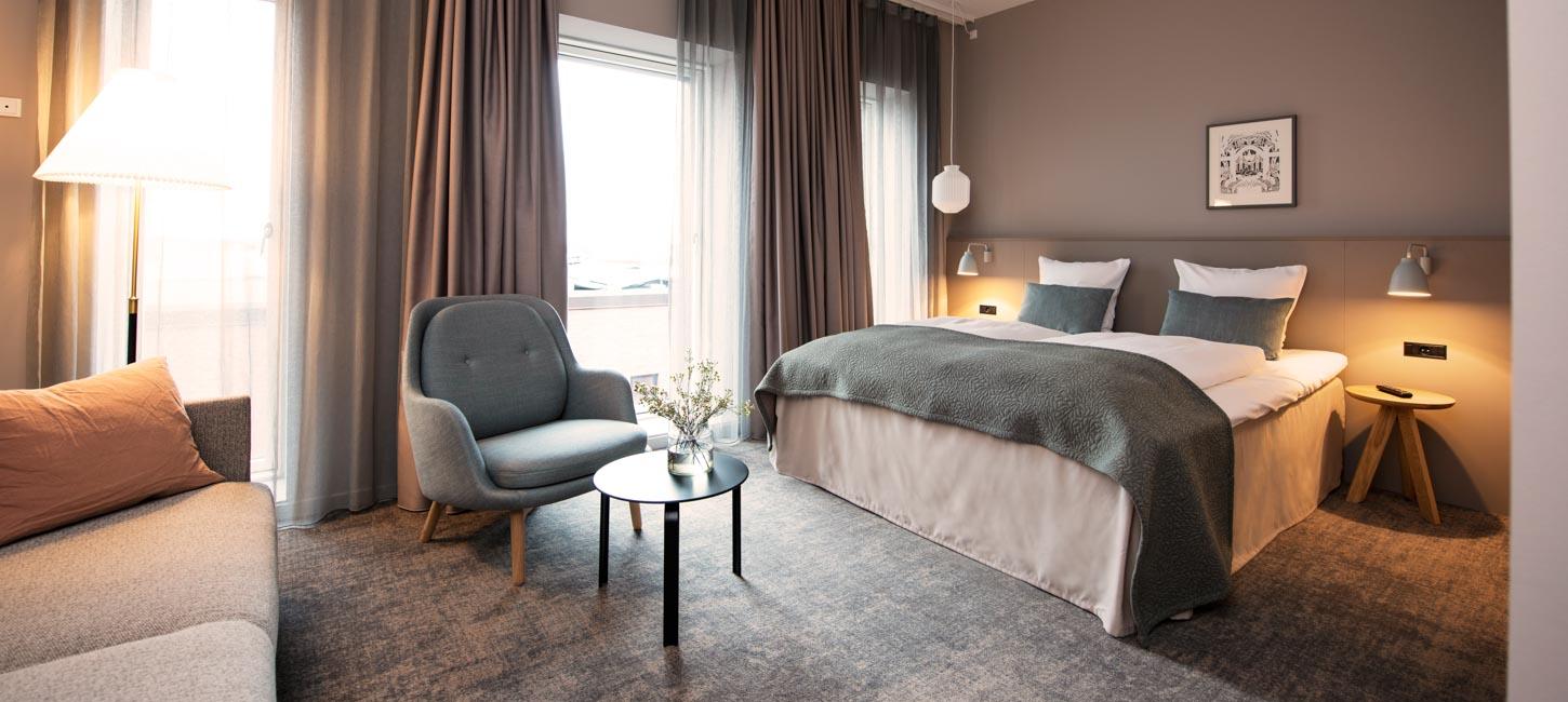Et hotelværelse med brune vægge og et gråt gulvtæppe. Til venstre ser man hjørnet af en grå sofa, og en læne stol med et lille sofabord til. Til højre står en dobbelt seng med hvide lagner og blå pyntepuder. På hver side står der et natbord og en natlampe.