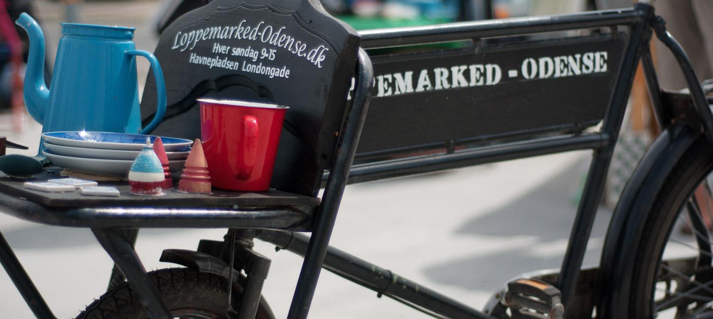 En nært billede af en sort gammel cykel. Cyklen har et lille lad foran med teksten "Loppemarked-Odense.dk" skrevet på. På ladet står en blå kande, par blå tallerkner og en rød kop.