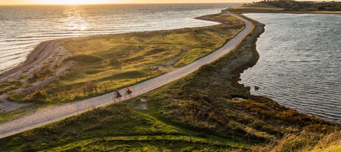 To cyklister cykler på smal grussti lige ud til vandet ved en fynsk strand. I baggrunden ses en smuk solnedgang. 