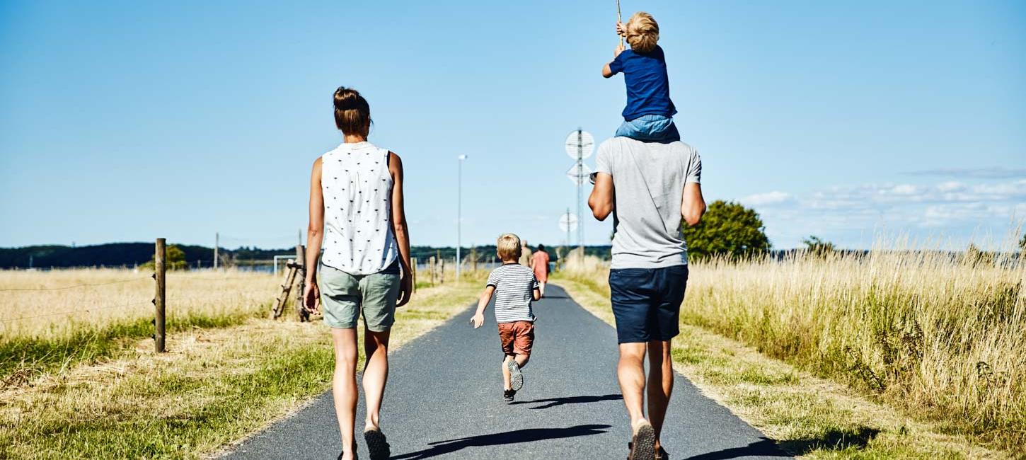En familie på fire går ned ad en landevej på en solrig dag. Manden til højre bærer et lille barn på sine skuldre. Mellem dem går et andet barn. De er omgivet af åbne marker, og himlen er klar og blå.