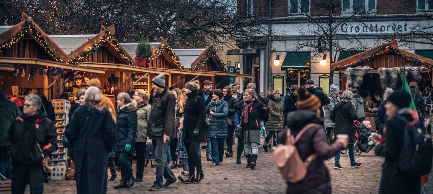 En brostensplads er fyldt med mennesker pakket ind i vintertøj. Rundt om pladsen står små træhytter pyntet med gran og julelys. Træhytterne har en åben front som giver adgang til de juleting de sælger.