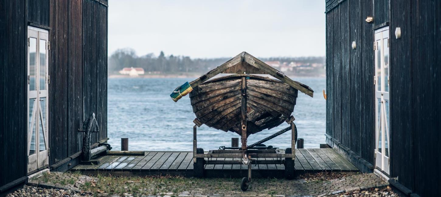 Mellem to sorte træværkshuse er en båd placeret på et stativ på havnen med udsigt til vandet. 