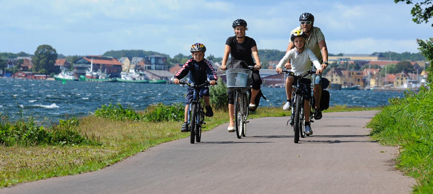 En familie på fire cykler sammen langs en sti ved kysten. Børnene fører an, og de voksne følger efter. I baggrunden ses det blå vand med en by helt i baggrunden. Det er en klar dag med solskin.