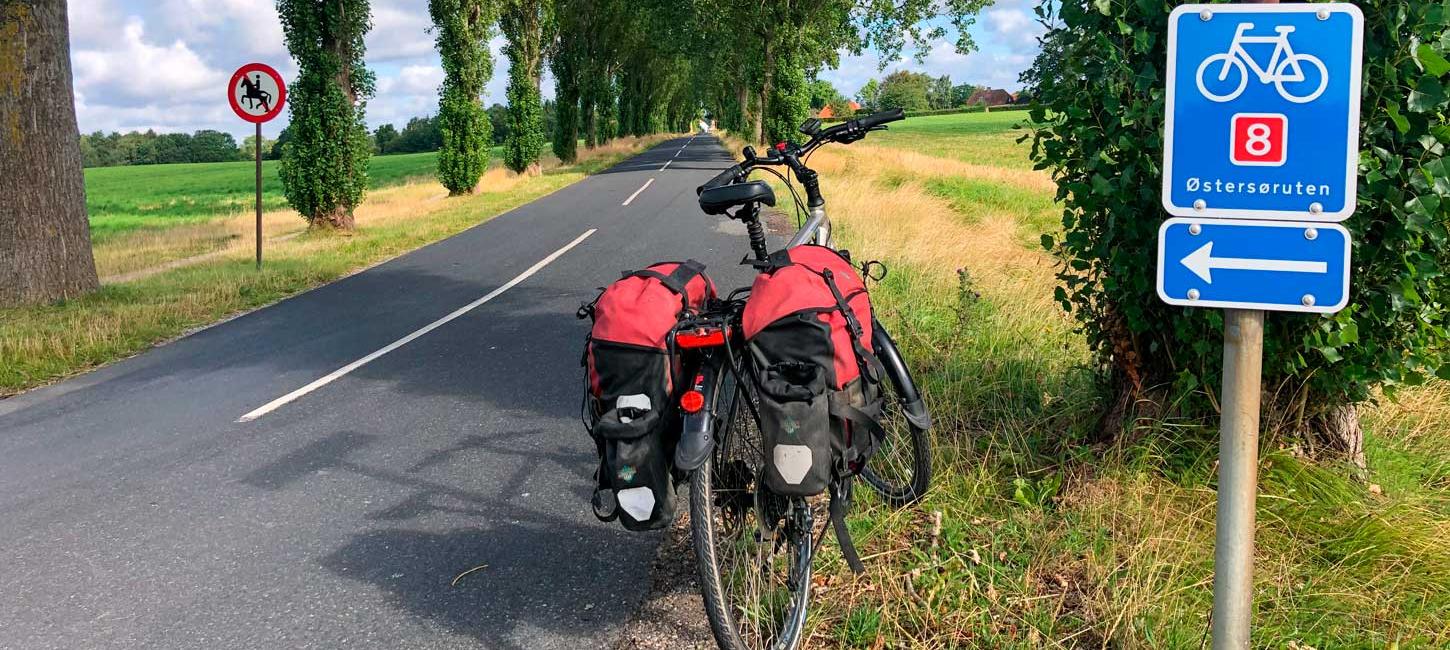Cykel med oppakning holder i vejkanten. I forgrunden ses et vejskilt, der viser vej på cykelruten Østersøruten. Det er sommer og solskin