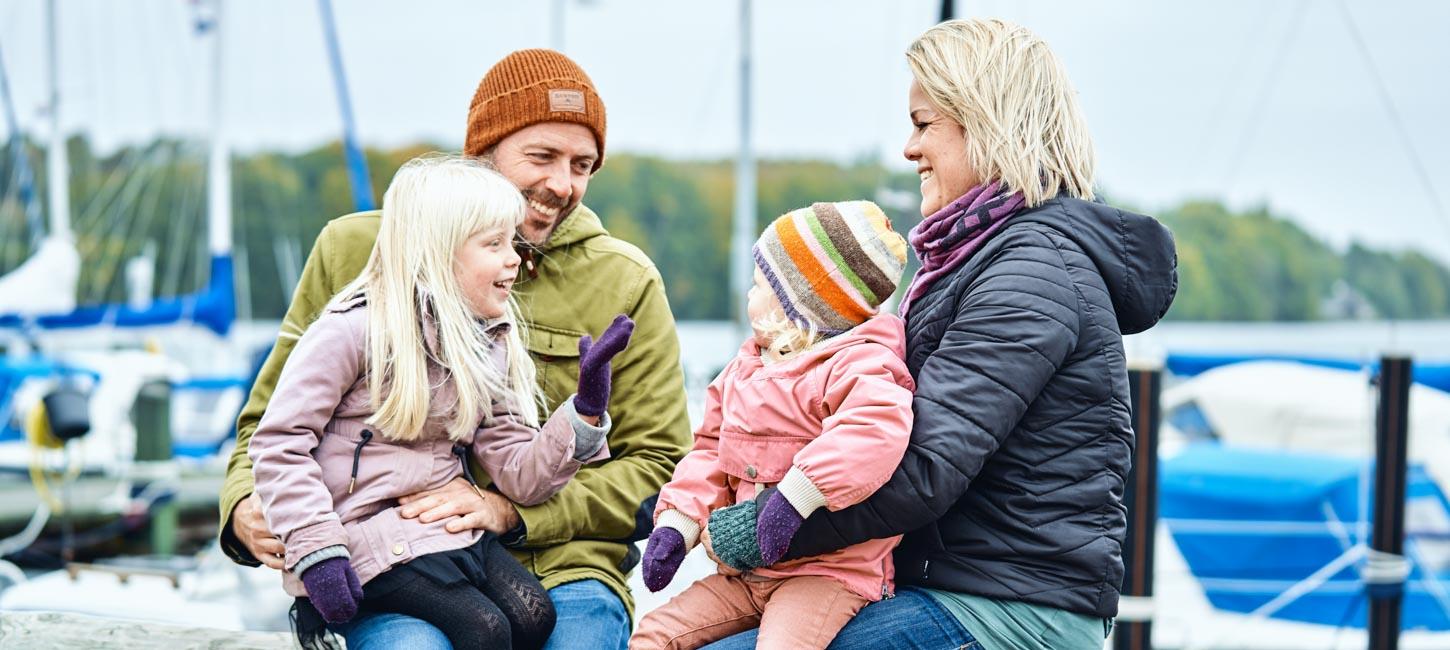 En mand og en dame sidder med hvert sit barn på skødet og i baggrunden er en havn