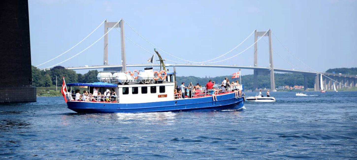 På havet foran Lillebæltsbroen sejler et blåt træskib med hvidt førerhus. På dækket står en flok mennesker og kigger udover vandet, og i bagenden flagre det danske flag.