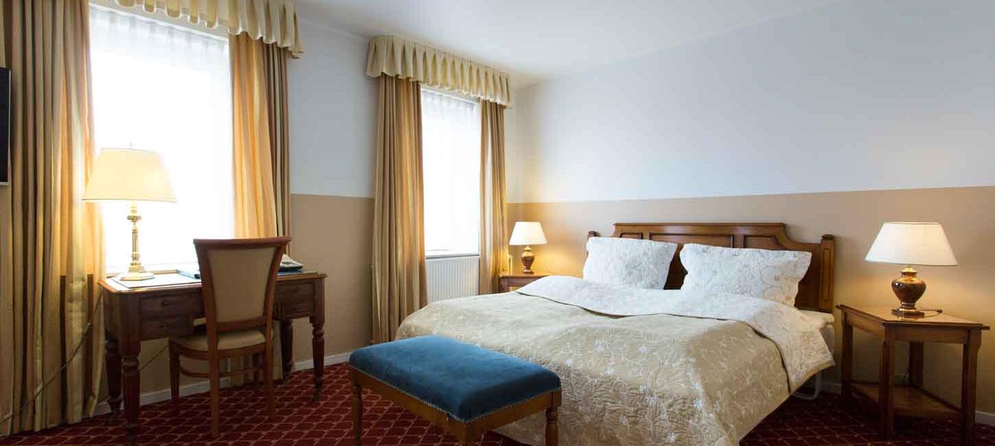 Et hotelværelse med en redt dobbeltseng, tændte lamper på bordene ved siden af sengen og et skrivebord foran et af vinduerne.