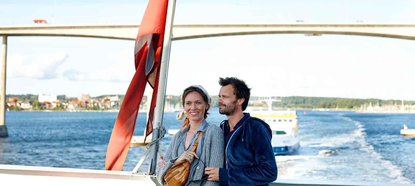Par står og holder om hinanden på den bagerste del af en færge og nyder udsigten. I baggrunden er Svendborgsund broen. 