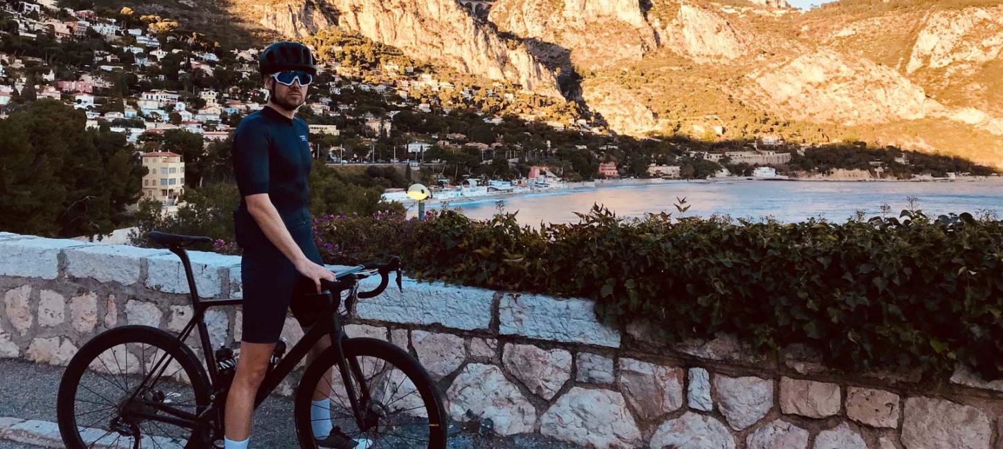 Cyklist i fuld cykel-outfit stående med sin cykel ved stengærde med sydlandsk landsby og hav i baggrunden.