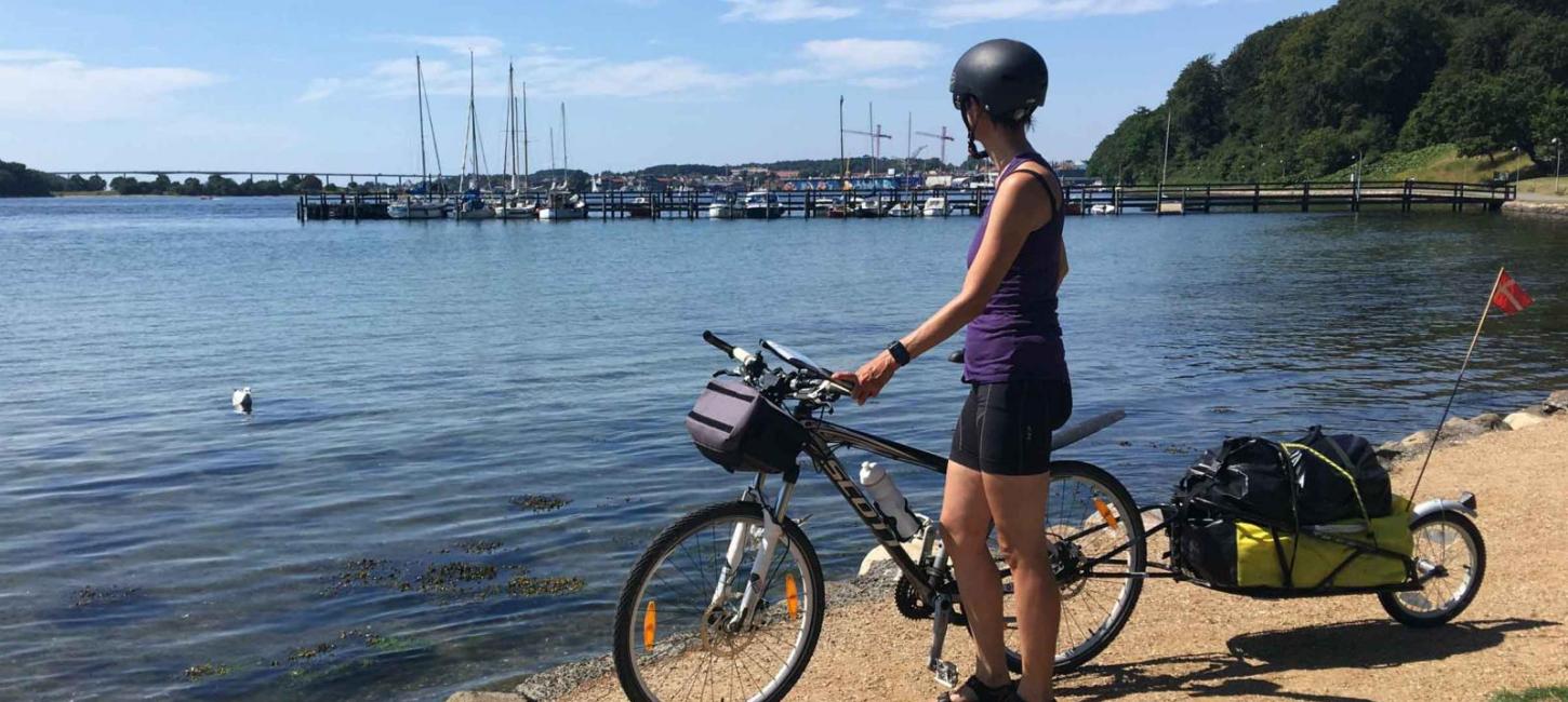 En kvinde står på en sti med sin cykel. cykelhjelm og cykeltrailer.  Hun kigger udover havet og havnen der er i baggrunden af billedet.
