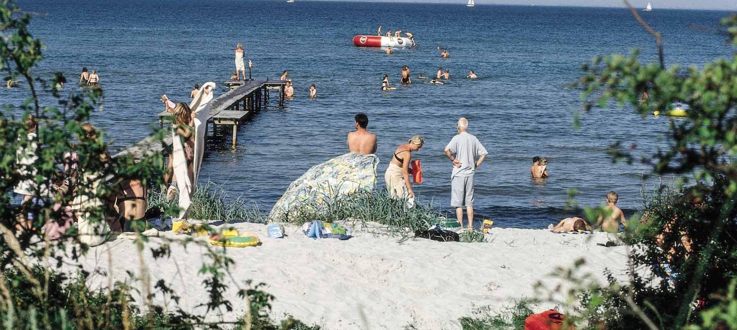 Strand med bølgende vand, badebro og mange badende gæster.