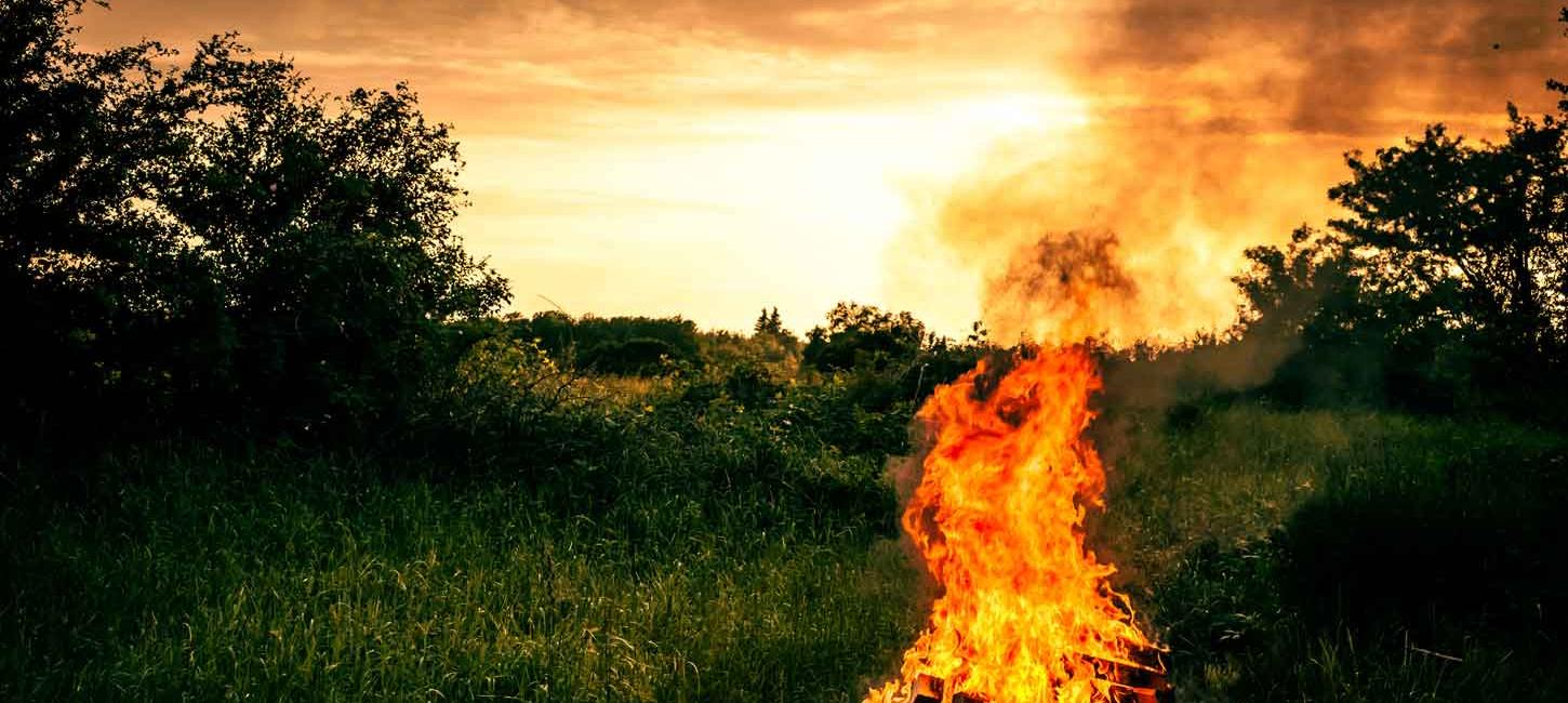 Orange solnedgang og mørkegrønne omgivelser med et stort Sankt Hans-bål i forgrunden.