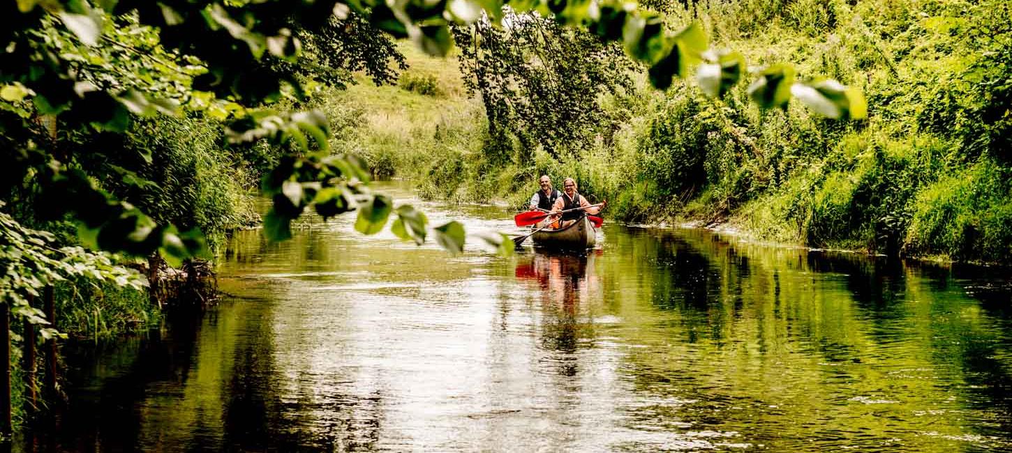En å er omgivet af grønt. Grønne buske og grønne træer hænger indover åen. På åen sejler et par en kano. De sidder i sorte redningsveste.