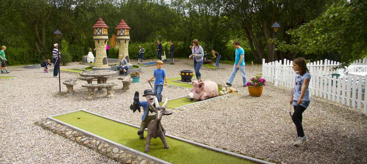 Minigolfbane med folk, der er i gang med at spille. På banen i forgrunden ses en figur af Klodshans på gedebukken.