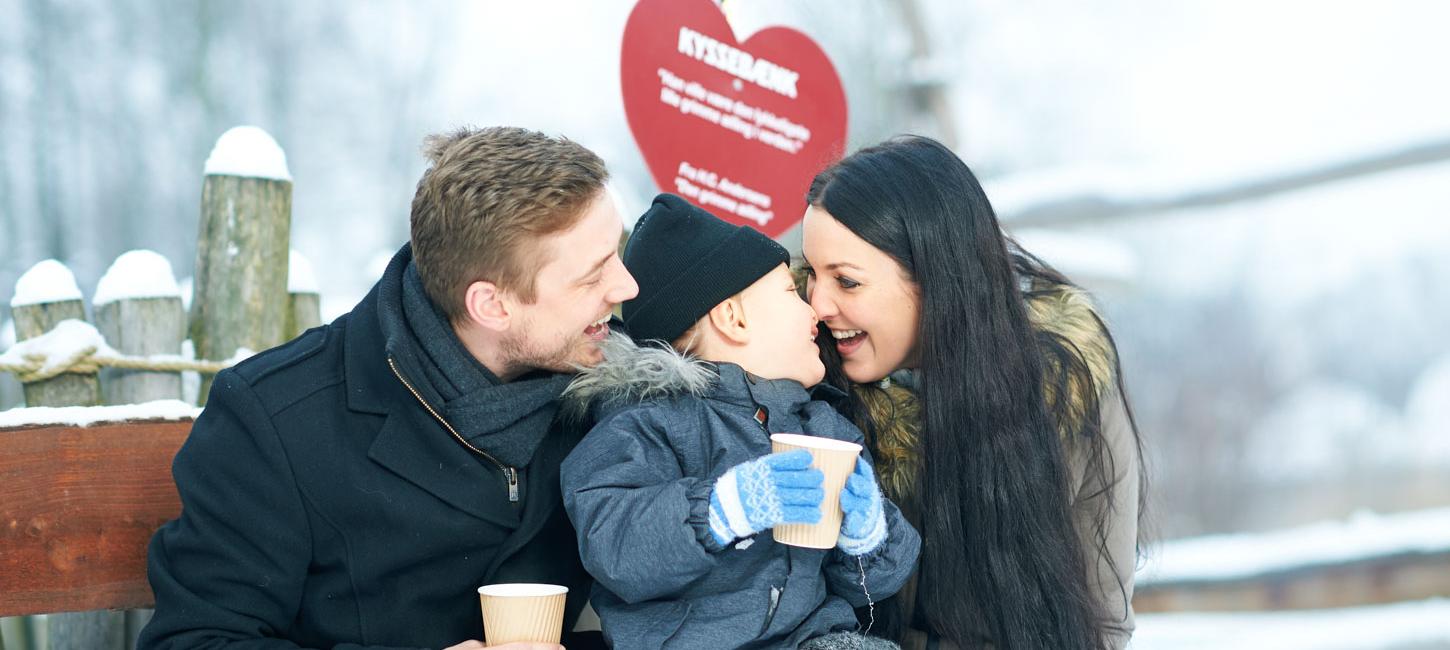 En mand og en kvinde sidder på en 'Kyssebænk' og griner. Imellem dem sidder en lille dreng. Der er sne omkring dem og de er alle klædt godt på.