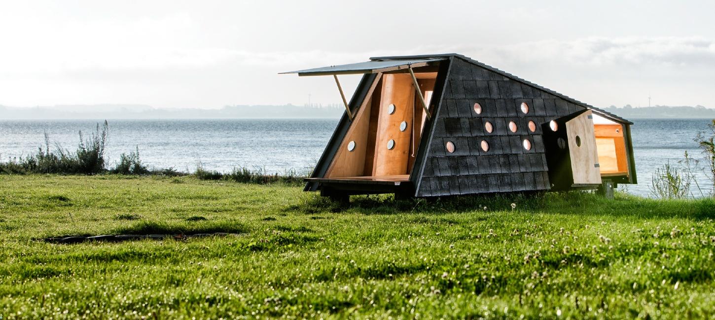 Shelter med åbne døre, der afslører et varmt interiør i træ. Det befinder sig i grønne omgivelser med udsigt over havet.