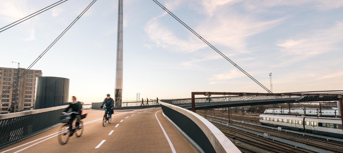 Byens Bro i Odense med to cyklister i fart på vej henover broen. Etagebygning i baggrunden, og togene nede på skinnerne til højre i billedet.