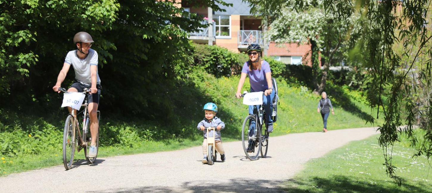 En mand, en kvinde og et lille barn på løbecykel cykler hen ad en solbeskinnet sti i grønne omgivelser.